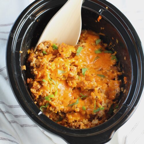 Crockpot insert on counter with Ground Pork Slow Cooker Casserole inside and melted cheese on top with scallions garnish and a wood spoon in it.