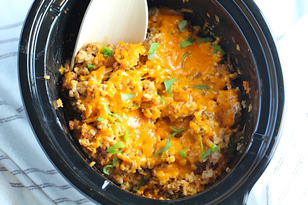 Crockpot insert on counter with Ground Pork Slow Cooker Casserole inside and melted cheese on top with scallions garnish and a wood spoon in it.