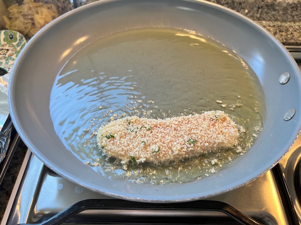 One strip of panko chicken starting to fry in shallow oil in a frying pan on the stove.