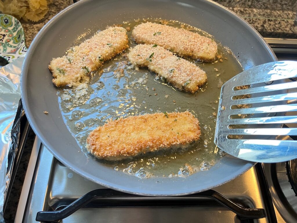Four pieces of panko chicken frying in shallow oil in a frying pan on the stove. Spatula has flipped one strip over and it's golden brown.