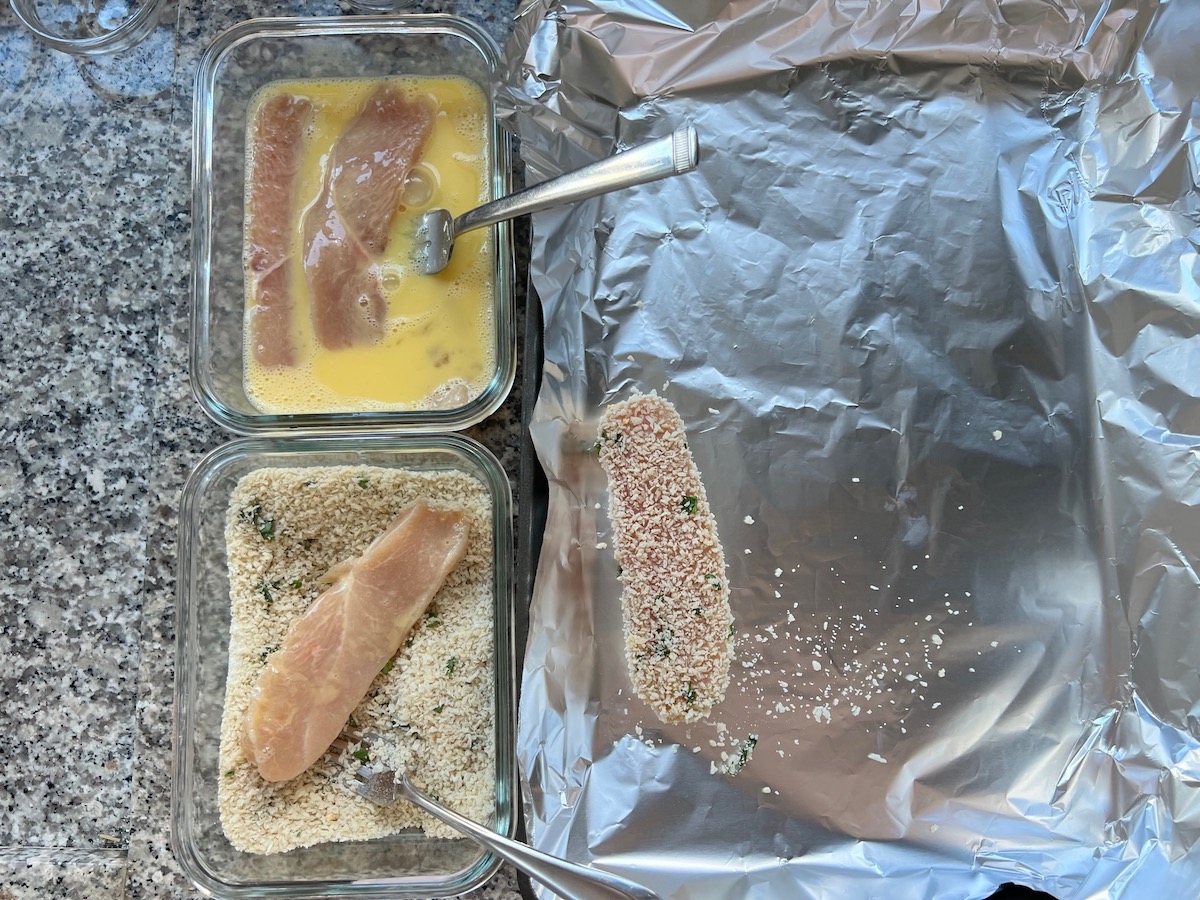 Raw chicken in a bowl of beaten eggs, below that, a bowl with panko breadcrumbs and a chicken strip with egg coating being breaded. Then to the right, a pan lined with aluminum foil with raw breaded Panko Chicken.