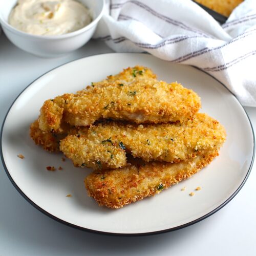 Several crispy and golden brown Panko Chicken strips stacked on a plate with a small bowl of Lime aioli off to the side.