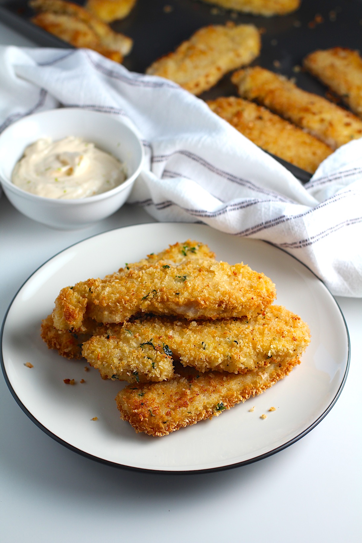 Several crispy and golden brown Panko Chicken strips stacked on a plate with a small bowl of Lime aioli off to the side and a sheet pan with more panko chicken pieces in background.