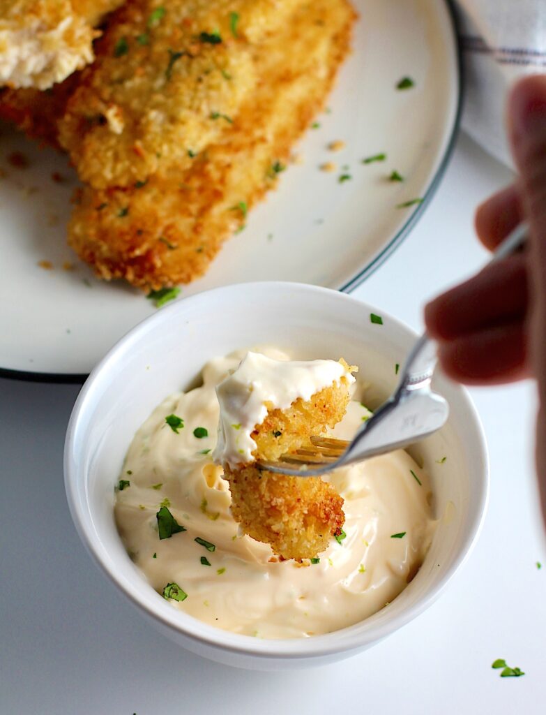 Fork dipping panko chicken into lime aioli sauce. Plate of more panko chicken strips in the background.