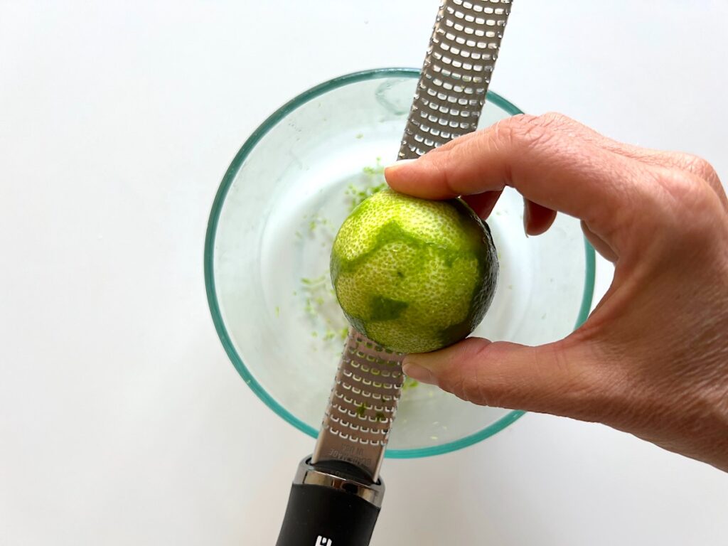 Hand zesting a lime on a microplane over a clear bowl for Cake with Lime recipe.
