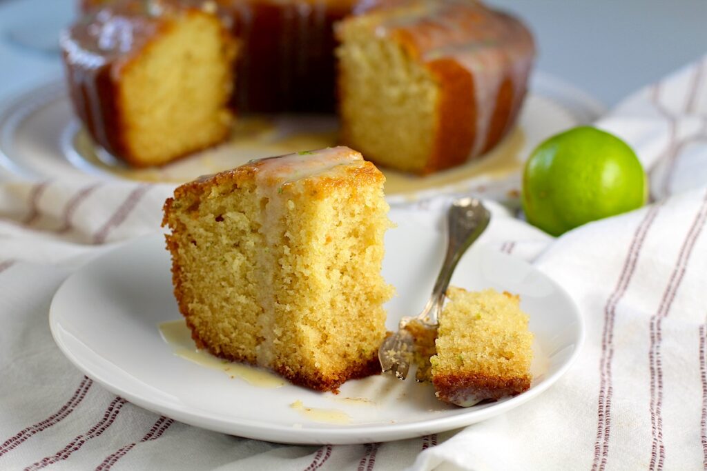 Moist Vanilla Cake with Lime slice standing on a plate with a fork that cut a bite on the plate. A green lime behind the plate and the rest of the bundt cake on a platter in the background.