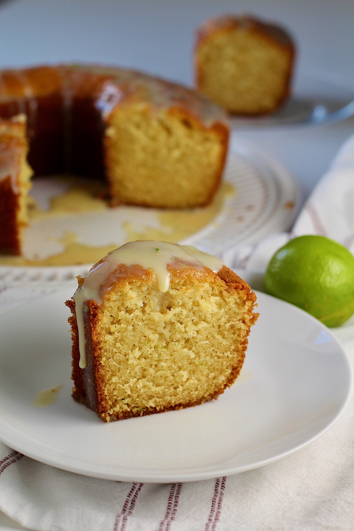 Moist Vanilla Cake with Lime slice standing on a plate with lime glaze dripping down.  A green lime behind the plate and the rest of the bundt cake on a platter in the background.