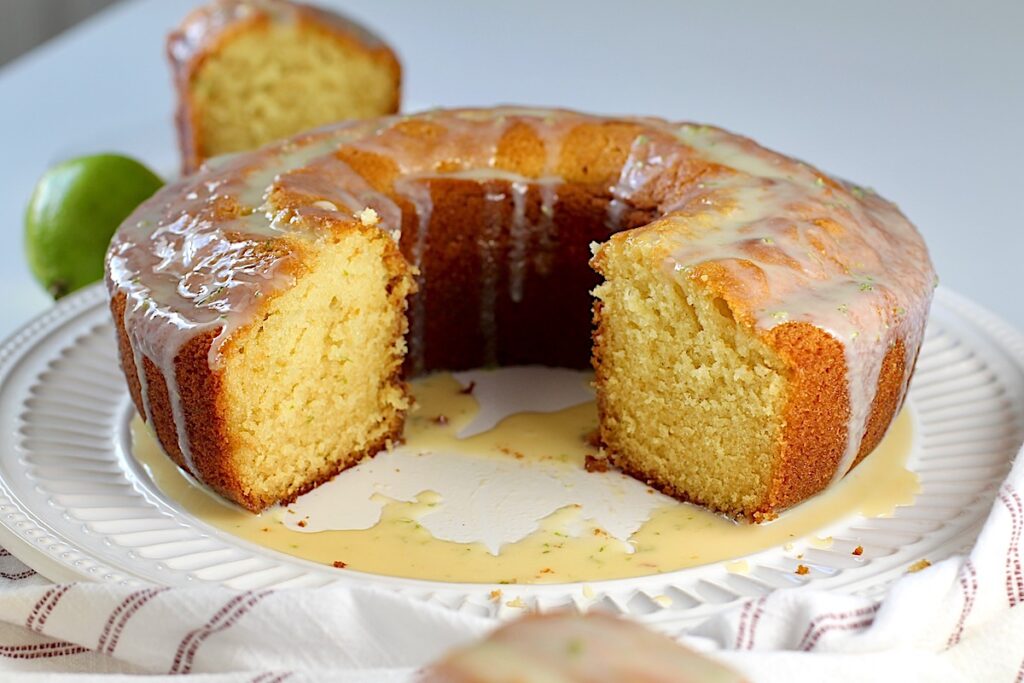 Vanilla bundt cake with lime on a platter with glaze poured over top and dropping on to the platter. A large section of the cake is missing from the front exposing the inside of the bundt cake.