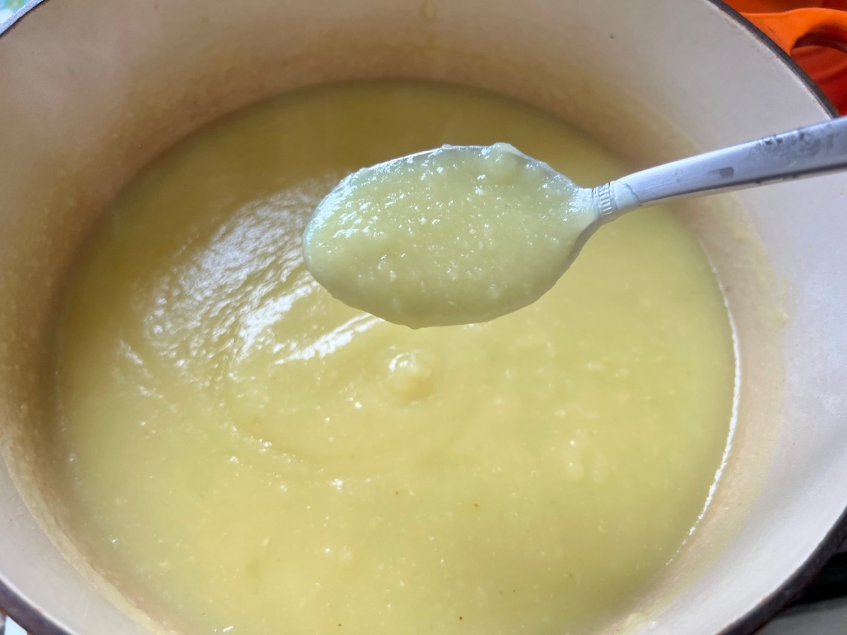Spoon scooping up pureed potato soup for Chicken Kale Soup thickened with pureed potatoes (Caldo Verde com Frango) in a pot on stove.