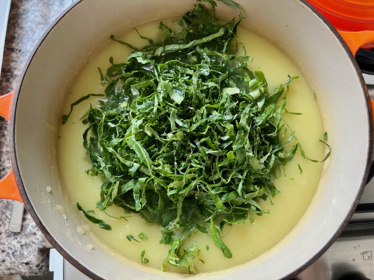 Shredded kale added to pureed potato soup for Chicken Kale Soup thickened with pureed potatoes (Caldo Verde com Frango) in a pot on stove.