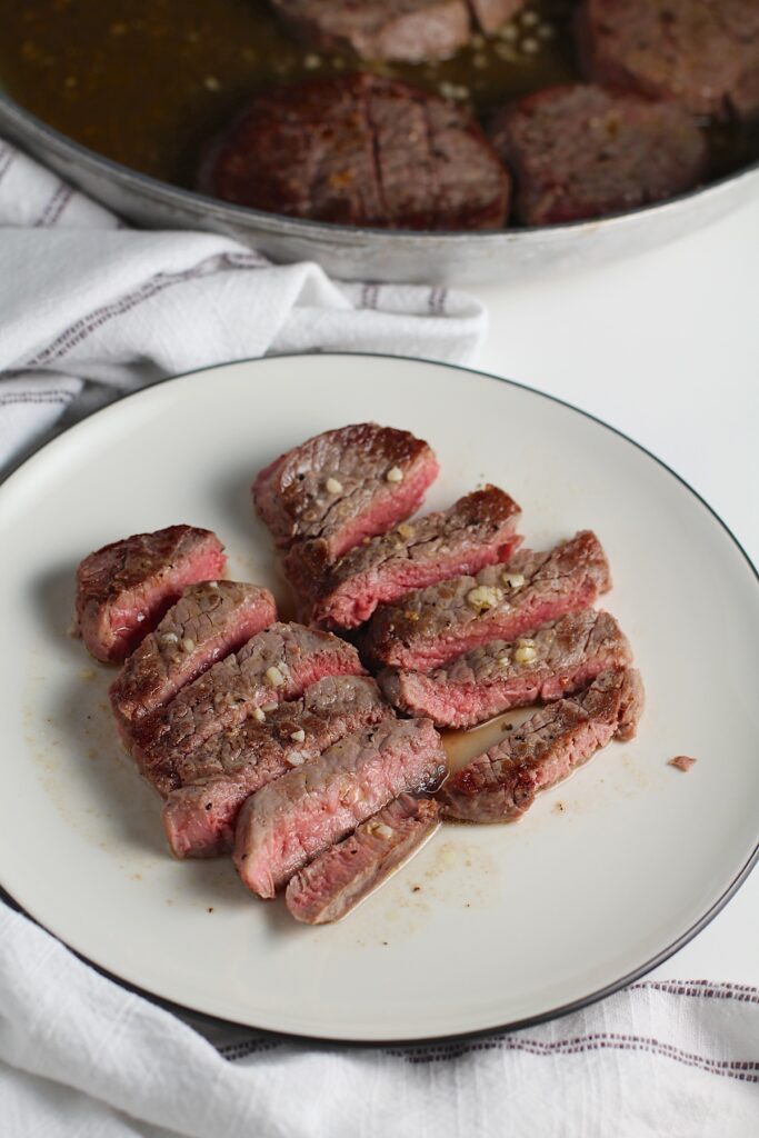 Sliced Filet of beef with garlic sauce on a plate with skillet with more beef in background.