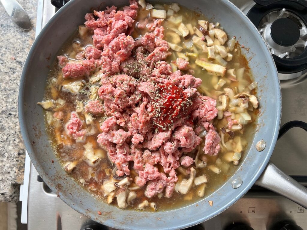 Broth, sausage, tomato paste, oregano added to cooked mushrooms, onions, and garlic for ragu for Spicy Sausage Pasta recipe.