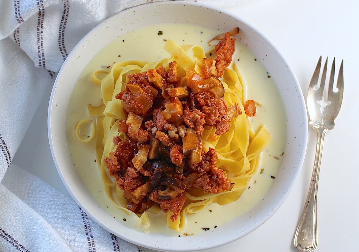 Spicy Sausage Pasta on a plate with Parmesan cream sauce on the bottom, fettuccine pasta on top of the sauce, and then Sausage and Mushroom ragu on top of the pasta.