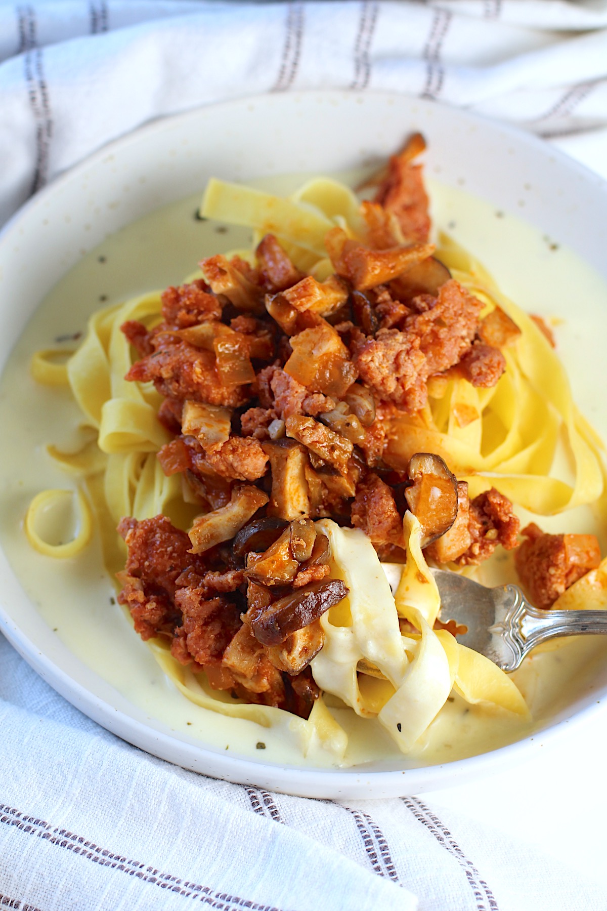 Spicy Sausage Pasta on a plate with Parmesan cream sauce on the bottom, fettuccine pasta on top of the sauce, and then Sausage and Mushroom ragu on top of the pasta.  A fork is scooping up pasta in the cream sauce. 