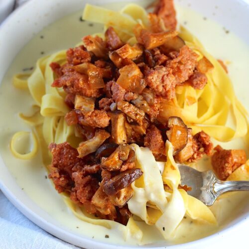 Spicy Sausage Pasta on a plate with Parmesan cream sauce on the bottom, fettuccine pasta on top of the sauce, and then Sausage and Mushroom ragu on top of the pasta. A fork is scooping up pasta in the cream sauce.