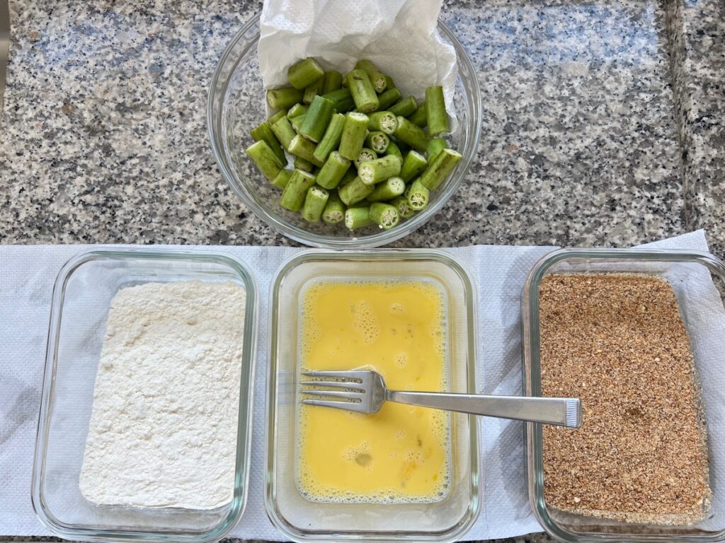 Cleaned and cut okra in a bowl next to 3 bowls: one with flour, one with beaten egg, and one with breadcrumb mixture for Breaded Okra in Air Fryer recipe.
