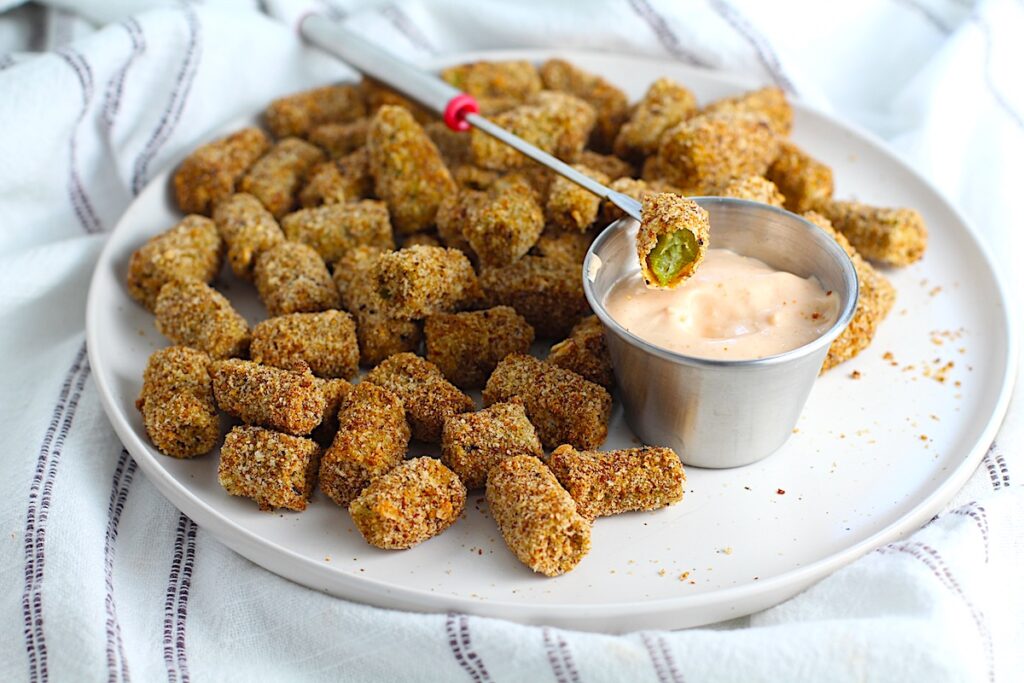 Pick holding a crispy golden breaded Okra with a bite taken out over a dipping sauce for Breaded Okra in air fryer recipe.