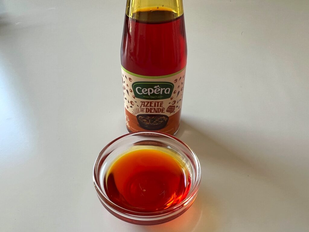 Small clear bowl with orange colored Dende oil and a bottle of dende oil in the background for Honey Lemon Pepper Chicken recipe.