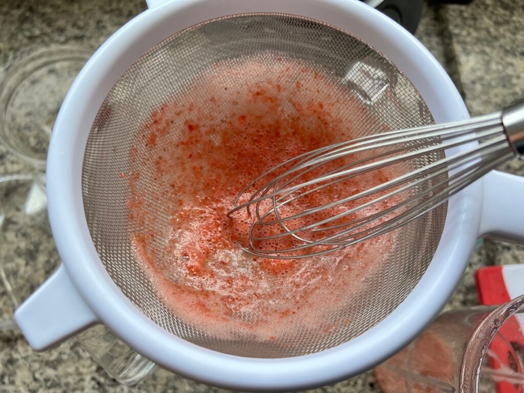Blended strawberries and tea going through a strainer to remove some of the seeds as it's added to the serving pitcher for how to make Strawberry Acai Refresher.