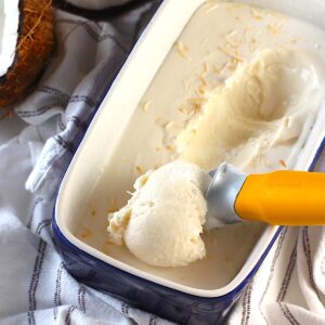 Ice Cream scoop scooping Coconut Ice Cream Recipe without Cream with toasted coconut on top in a blue loaf pan. In the background is a fresh coconut cracked in half.