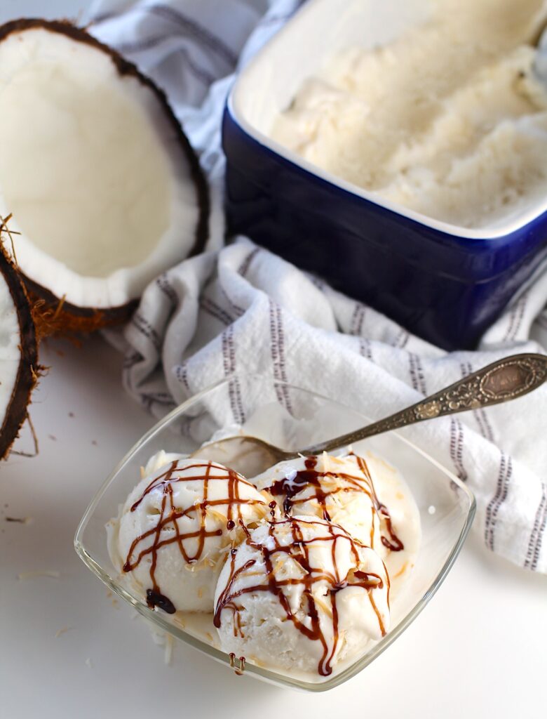 Three scoops of Coconut Ice Cream Recipe without Cream in a clear bowl with chocolate sauce drizzled on top and spoon in bowl. In the background is a the rest of the ice cream in blue loaf pan and a fresh coconut cracked in half.