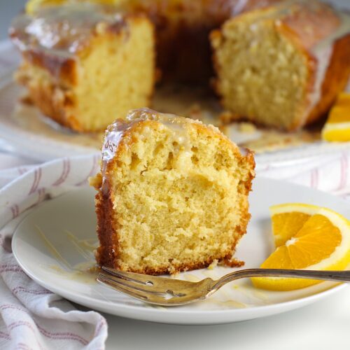 Piece of Orange Bundt Cake (Bolo de Laranja) on a plate with the rest of the cake in background. There are orange slices on the side of the plate and a fork on the plate.