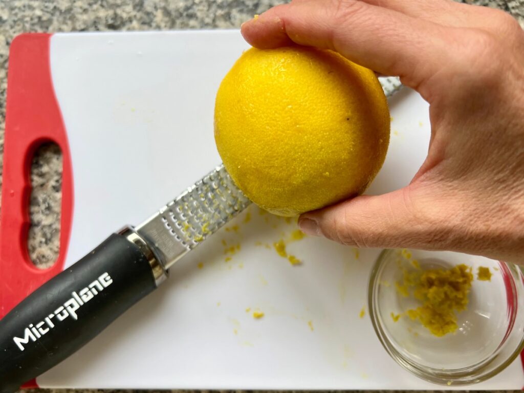 Hand zesting an orange over a microplane on a cutting boars with a bowl of zest off to the side for Orange Bundt Cake recipe.
