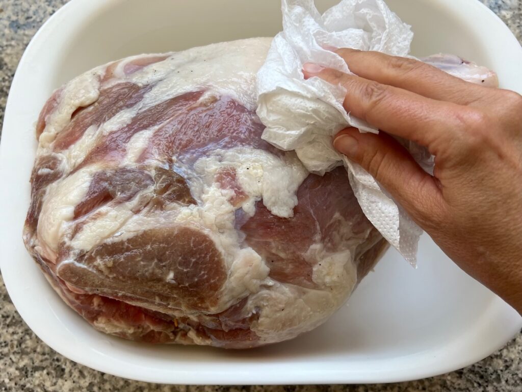 Hand patting a pork shoulder with bone in dry in a large pan after rinsing the brine off.