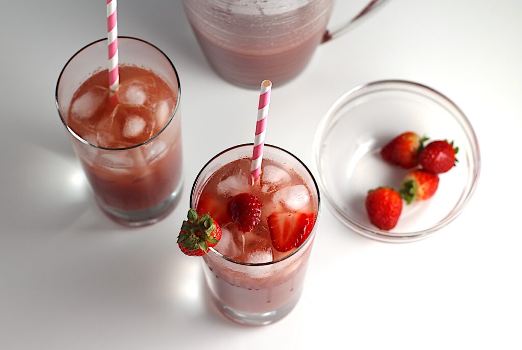 Two glasses of Strawberry Acai drink with pink and white striped straws and strawberries in the front glass. In the background is the pitcher of the rest of the drink for this article How to make a strawberry acai refresher.