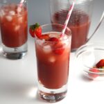 Two glasses of Strawberry Acai drink with pink and white striped straws and strawberries in the front glass. In the background is the pitcher of the rest of the drink for this article How to make a strawberry acai refresher.
