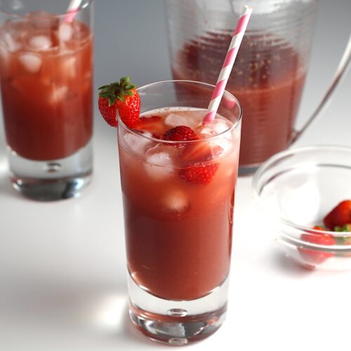 Two glasses of Strawberry Acai drink with pink and white striped straws and strawberries in the front glass. In the background is the pitcher of the rest of the drink for this article How to make a strawberry acai refresher.