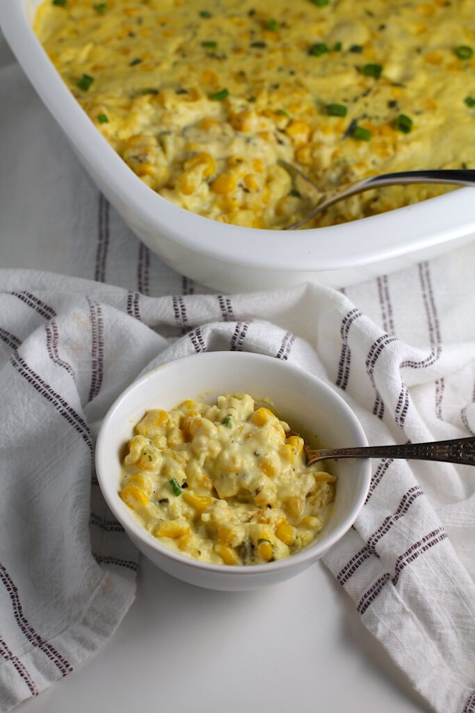 Corn Casserole Recipe with Cream Cheese and scallion slices on top in a bowl with spoon and casserole dish in background.