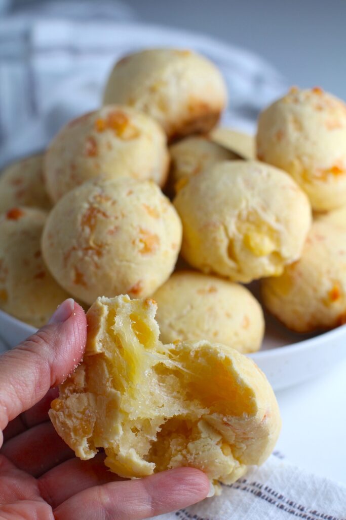 Hand ripping apart a gooey Gluten Free Brazilian Cheese bread, or Pao de Queijo, with a bowl stacked with more breads in the background.