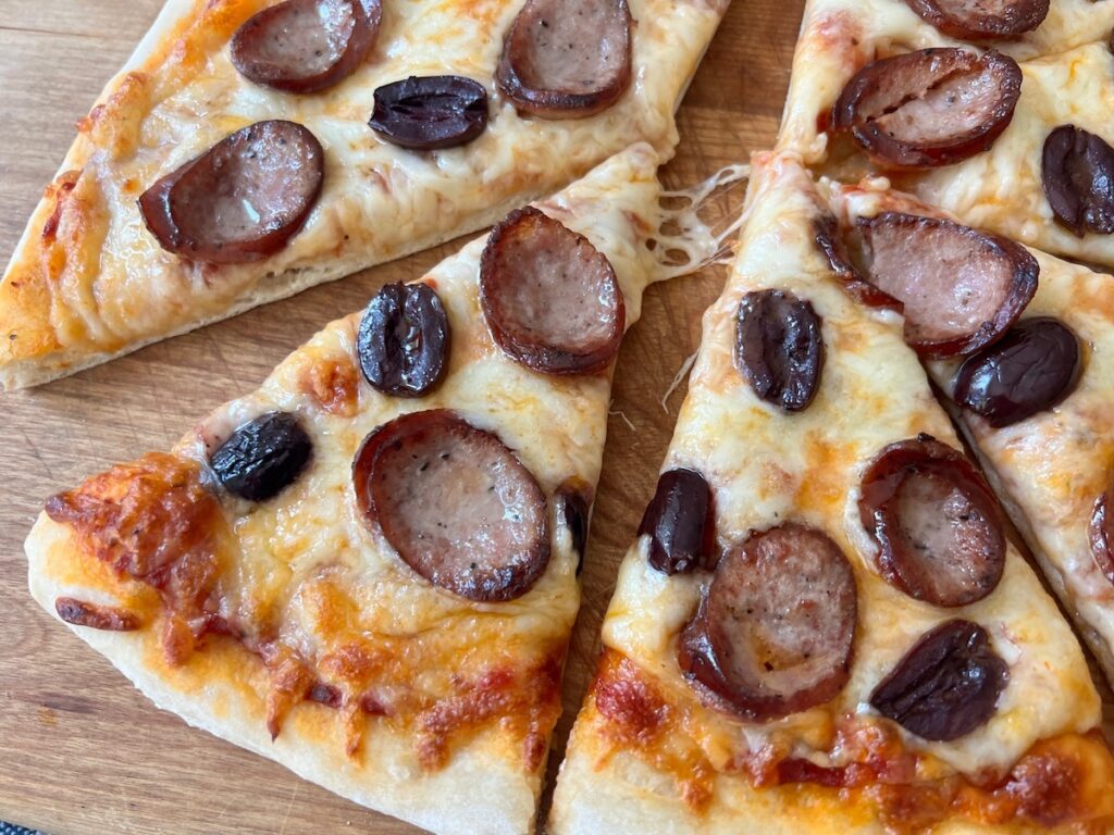 Brazilian Pizza cut into triangles on a wood cutting board on blue and white towel on table. It's a thin, crispy crust topped with mozzarella cheese, cabresa sausage, and olives.