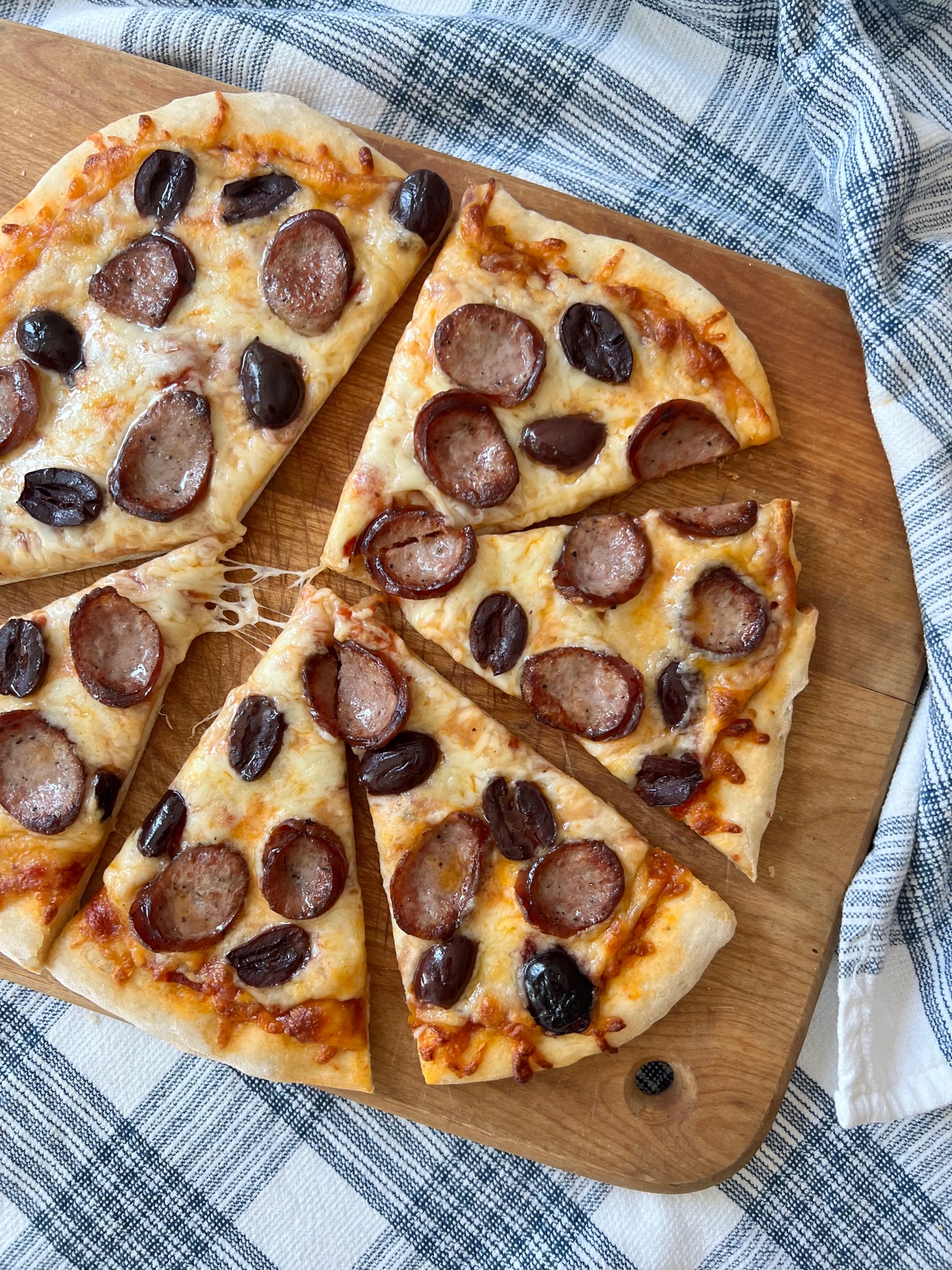 Brazilian Pizza cut into triangles on a wood cutting board on blue and white towel on table. It's a thin, crispy crust topped with mozzarella cheese, cabresa sausage, and olives.