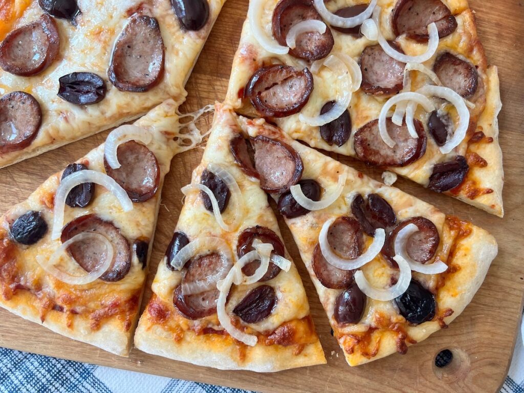 Brazilian Pizza cut into triangles on a wood cutting board on blue and white towel on table. It's a thin, crispy crust topped with mozzarella cheese, cabresa sausage, onions, and olives.