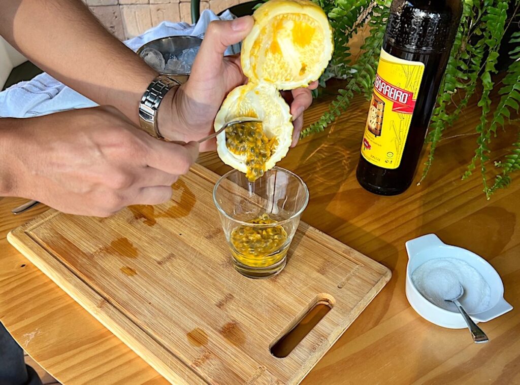 Hand holding a Yellow passion fruit cut in half with inside pulp being poured into a short martini glass on a cutting board, next to a bottle of cachaca and bowl of sugar for a Passion Fruit Caipirinha recipe.