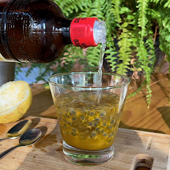 Bottle pouring cachaca into a martini glass with passion fruit pulp and sugar on a cutting board with cut yellow passion fruit to the left and a bottle of cachaca in background for a Passion Fruit Caipirinha recipe.