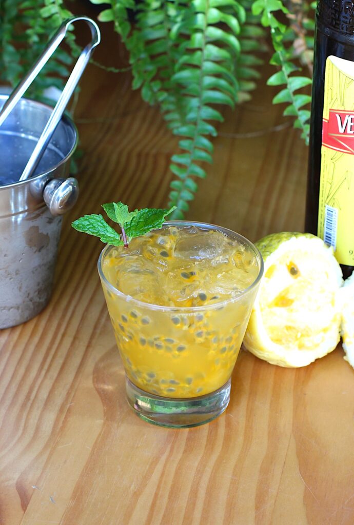 Passion Fruit Caipirinha in a martini glass on a cutting board with a bucket of ice to the left and a cut yellow passion fruit and a bottle of cachaca to the right in background.
