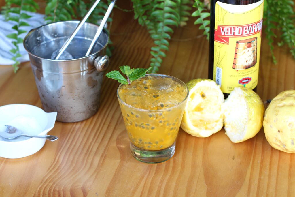 Passion Fruit Caipirinha in a martini glass on a cutting board with a bucket of ice to the left and a cut yellow passion fruit and a bottle of cachaca to the right in background.
