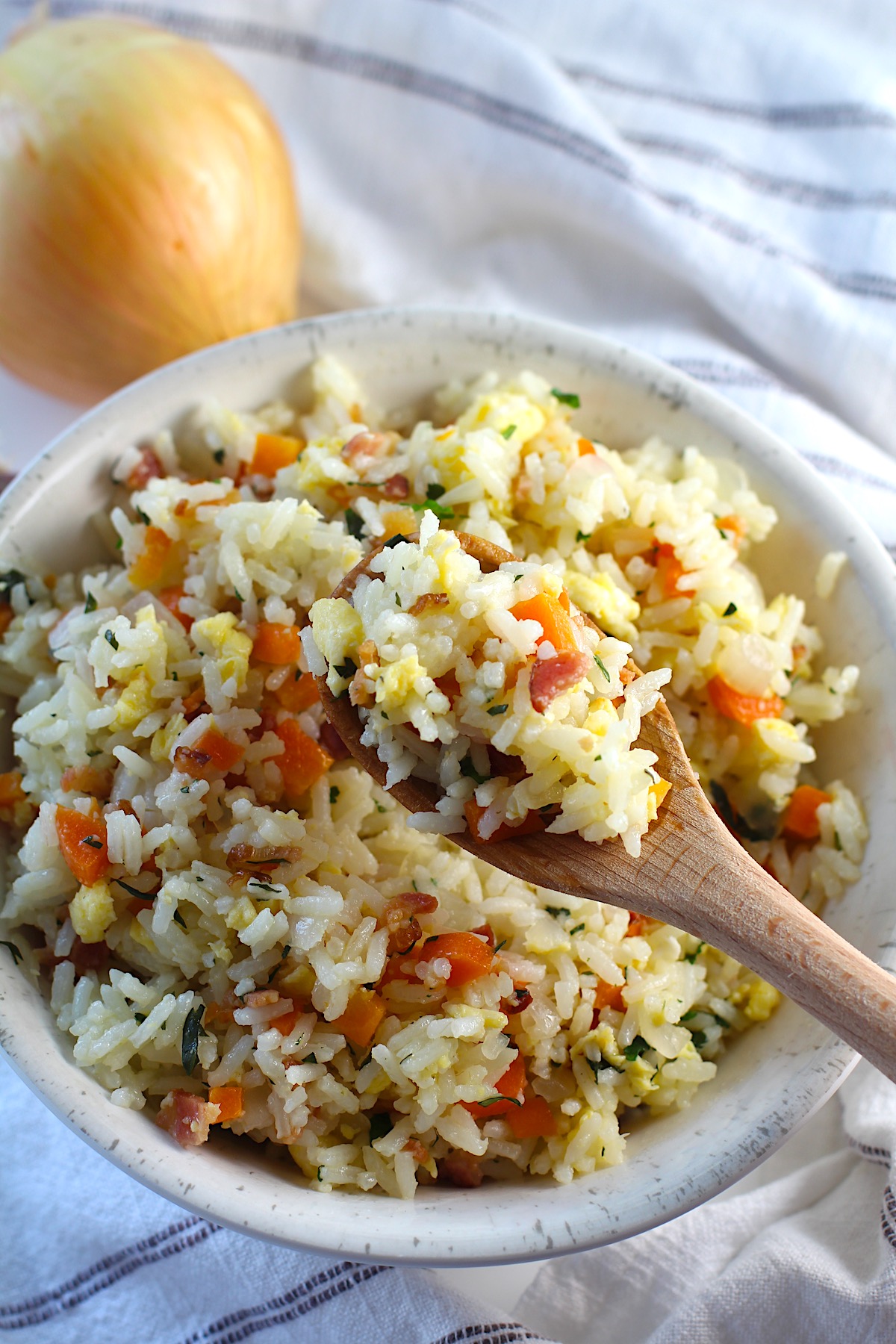 Arroz pilaf scoop in a wood spoon over a bowl of arroz pilaf with carrots, bacon, and cilantro mixed in and a spoon in the bowl.