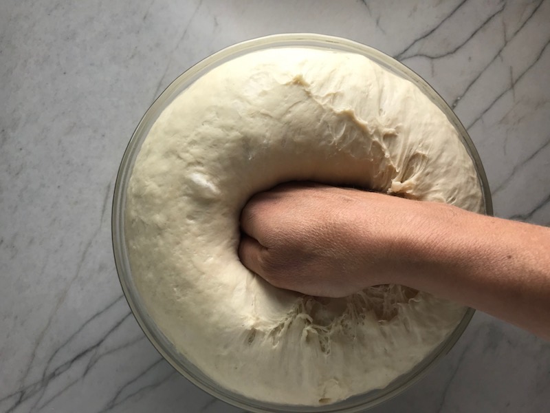 Hand punching down risen Brazilian Pizza dough in a glass bowl.