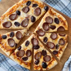 Brazilian Pizza cut into triangles on a wood cutting board on blue and white towel on table. It's a thin, crispy crust topped with mozzarella cheese, cabresa sausage, and olives.