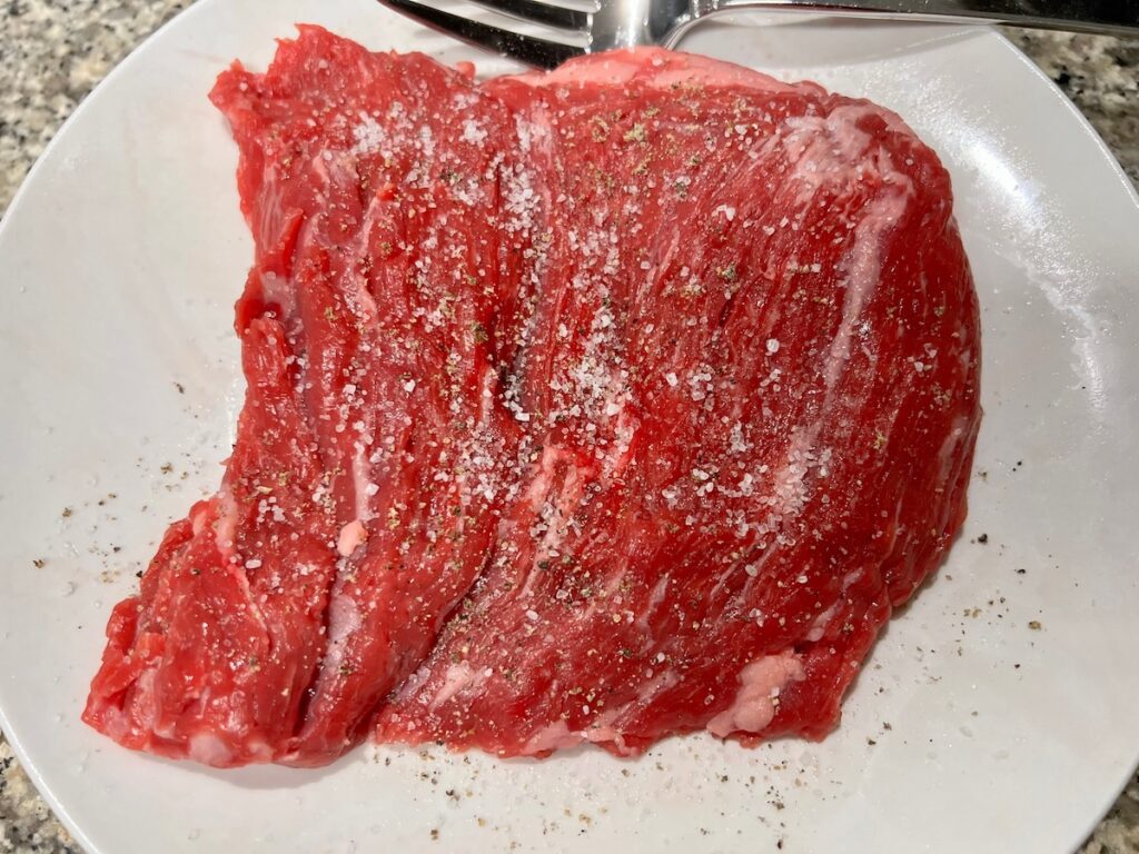 Raw flank steak on a plate with coarse salt and pepper all over it for Churrasco in Air Fryer.
