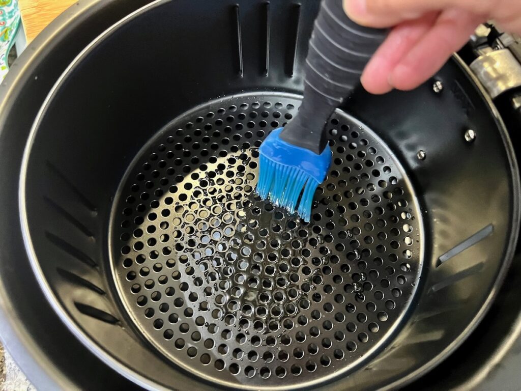 Hand using silicone brush to spread olive oil on the bottom of the air fryer basket.