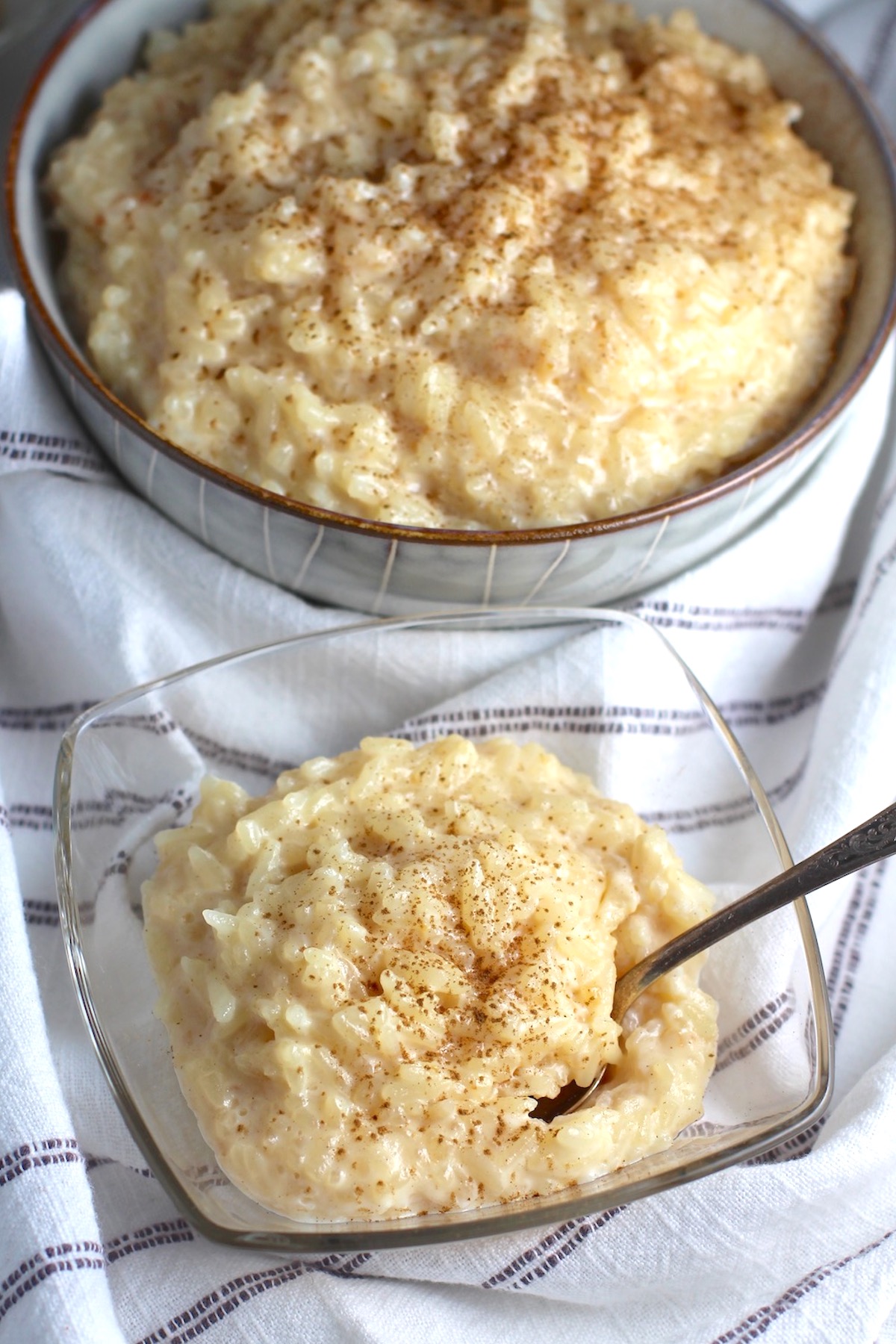 Rice Pudding with condensed milk in a clear serving bowl with cinnamon on top and a spoon scooping.  A larger bowl of rice pudding is in the background.