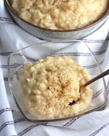 Rice Pudding with condensed milk in a clear serving bowl with cinnamon on top and a spoon scooping. A larger bowl of rice pudding is in the background.