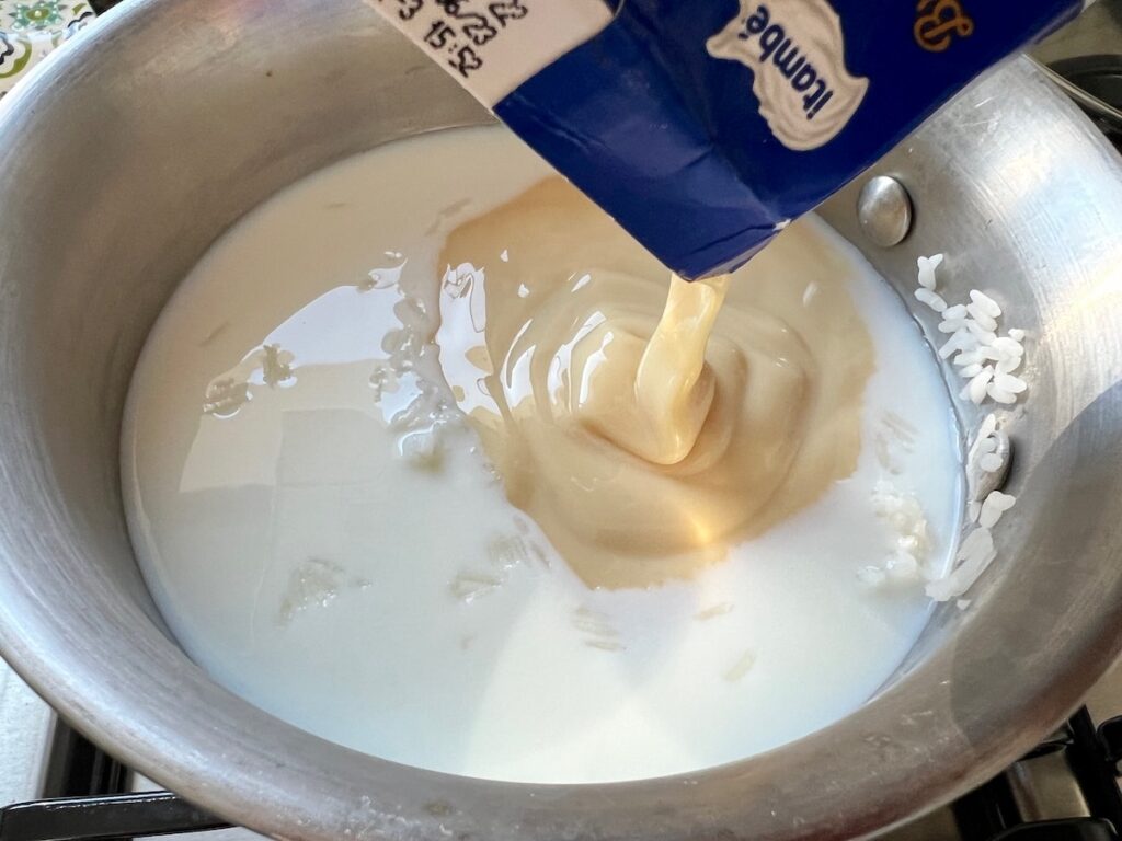 Condensed milk being poured into milk and uncooked white rice in a pot for Rice Pudding with condensed milk.