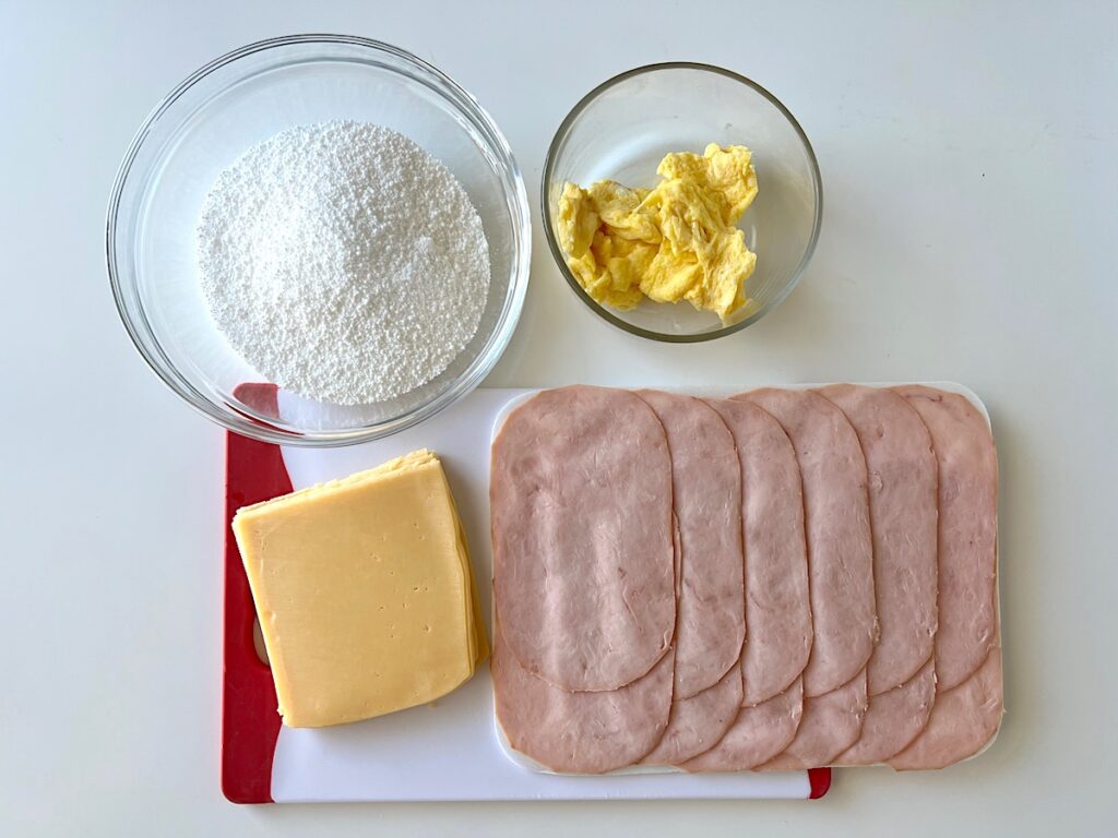 Bowl of tapioca starch, bowl of scrambled eggs, sliced ham, and sliced cheese all out on counter Brazilian Breakfast Tapioca.