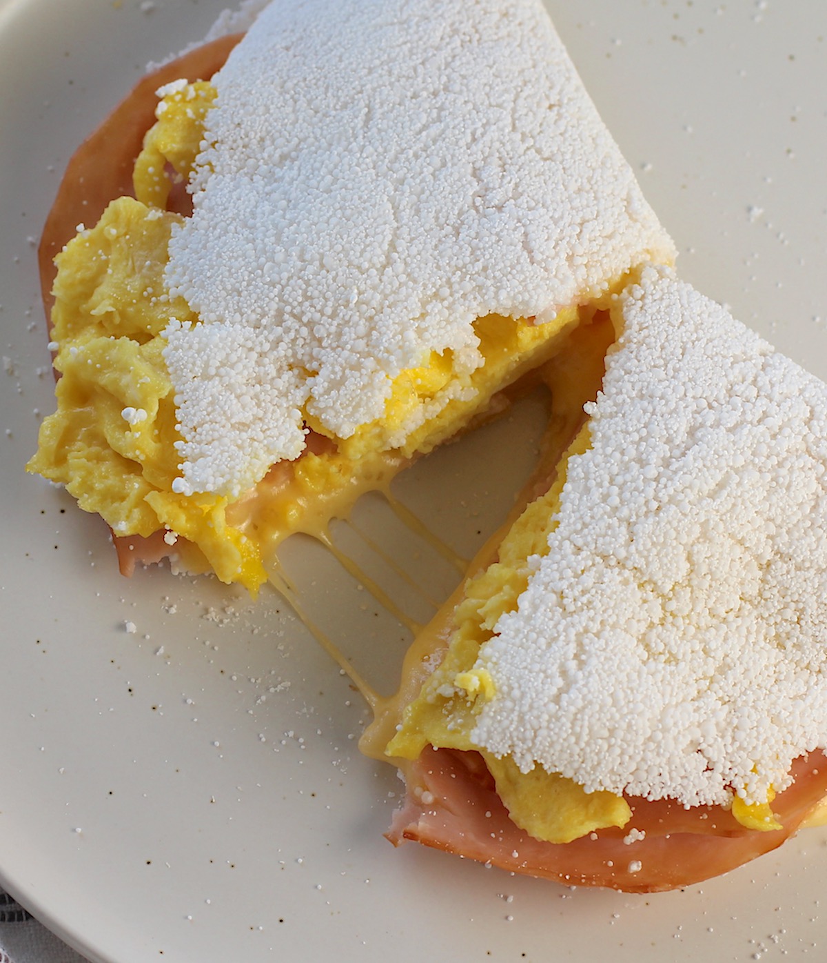 Brazilian Breakfast Tapioca crepe filled with ham, egg, and cheese cut in half with melted cheese pulling in the center on the plate. 